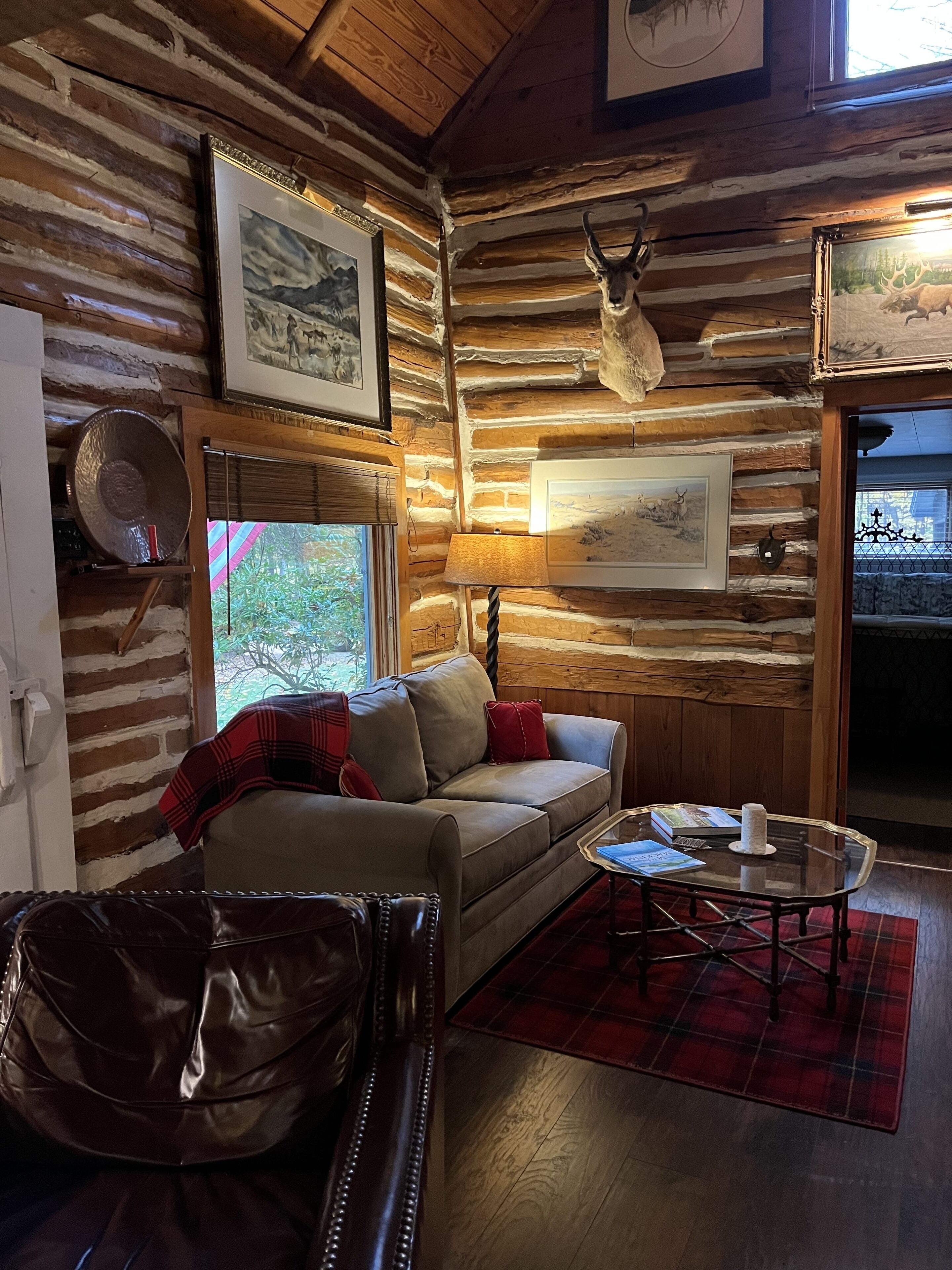 A cozy log cabin interior with a sofa, a plaid rug, a table with books and a candle, and framed artwork on the walls.