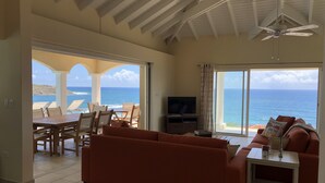 A spacious living room with a large sofa, a TV, and a dining area with chairs and a table. The room has a view of the ocean through large windows. - Luxury Villa w Spectacular Ocean Views, private pool/Hottub, Walk to Beach (Upper Prince's Quarter)