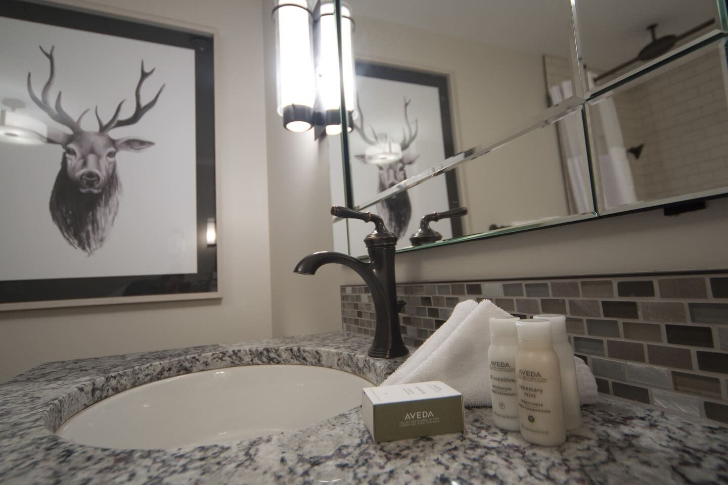 A bathroom with a granite countertop, a white sink, a framed deer head artwork, a mirror, and toiletries.