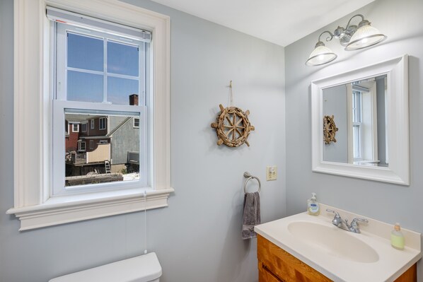 A bathroom with a large window, a ship's wheel decoration, a sink with a mirror, and a towel rack. - Plum Island Retreat-Beachside Stay Near Wildlife Refuge (Newbury)