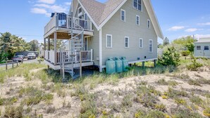 Ein zweistöckiges Haus mit Balkon und kleiner Terrasse, umgeben von Sand und vereinzelter Vegetation.