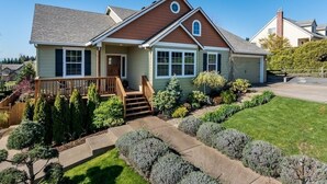 A two-story house with a front porch, a small deck, and a well-maintained lawn with various shrubs and a pathway leading to the front door.