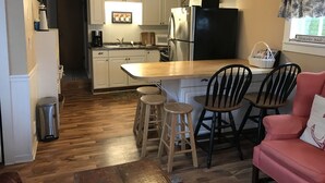 Een compacte keuken met witte kasten, een zwarte koelkast, een houten eettafel en twee barstoelen.