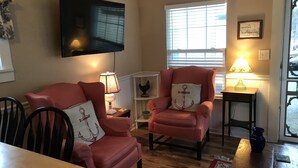 Een woonkamer met een houten eettafel, twee roze fauteuils, een aan de muur bevestigde televisie en een raam met jaloezieën.