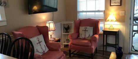 A living room with a wooden dining table, two pink armchairs, a television mounted on the wall, and a window with blinds.