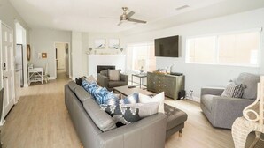 A spacious living room with a grey sofa, a grey armchair, and a coffee table. There is a fireplace, a TV mounted on the wall, and a ceiling fan. - Beautiful 6bd/7bath steps to the sand (Newport Beach)