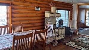 Interior de una cabaña de madera con una mesa de comedor, sillas, una estufa antigua y una ventana con persianas.