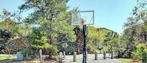 Canasta de baloncesto con una cerca de malla, árboles y un edificio al fondo.