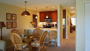 A modern kitchen with wooden cabinets, a central island, and a dining area with wicker chairs. - Enjoy a Piece of Paradise with Ocean View, Whale Watching, Snorkeling and More! (Kihei)