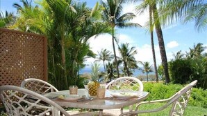 A round glass table with white wicker chairs, set outdoors with a view of palm trees and a body of water. - Enjoy a Piece of Paradise with Ocean View, Whale Watching, Snorkeling and More! (Kihei)