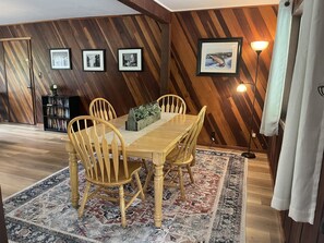 Uma área de refeições com uma mesa de madeira, quatro cadeiras, um tapete e uma estante.