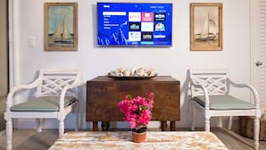 A living room with a flat-screen TV mounted on the wall, two white chairs with green cushions, a wooden coffee table with a potted plant, and a tray of seashells on a wooden side table.