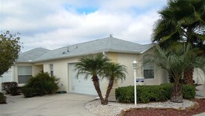 A single-story house with a garage, surrounded by a small garden with palm trees and shrubs. - Charming 3/2 Virginia Trace Villa, next to Lake Sumter Landing, with golf cart (The Villages)