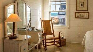 Ein Zimmer mit einem Holzschaukelstuhl, einem Schminktisch mit Glasplatte und einem Spiegel sowie einem Fenster mit Blick auf Strand und Gebäude.