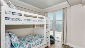 Un lit superposé avec une couvre-lit à motifs floraux, une fenêtre donnant sur la mer et un fauteuil en forme de poire.