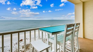 Un balcon avec une table et des chaises blanches, donnant sur une plage aux eaux d’un bleu limpide et au rivage sablonneux.
