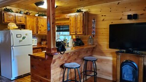 L’intérieur d’une cabane en bois comprend un coin cuisine équipé de meubles en bois, d’un réfrigérateur, d’un four à micro-ondes, d’une cuisinière, de deux tabourets de bar, d’une télévision et d’une cheminée.