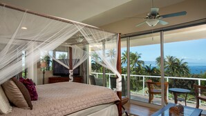 Une chambre à coucher avec un lit à baldaquin, un ventilateur de plafond et un balcon offrant une vue sur les palmiers et l’océan.