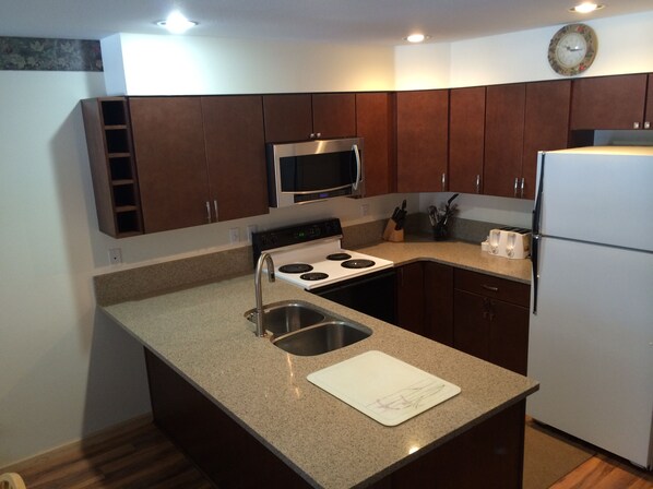 A compact kitchen with dark wooden cabinets, a stainless steel microwave, a white refrigerator, and a granite countertop.