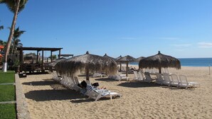 A beach with lounge chairs and thatched umbrellas, palm trees, and a clear sky. - Ground Floor, Oceanview And Oceanfront  (San Jose del Cabo)