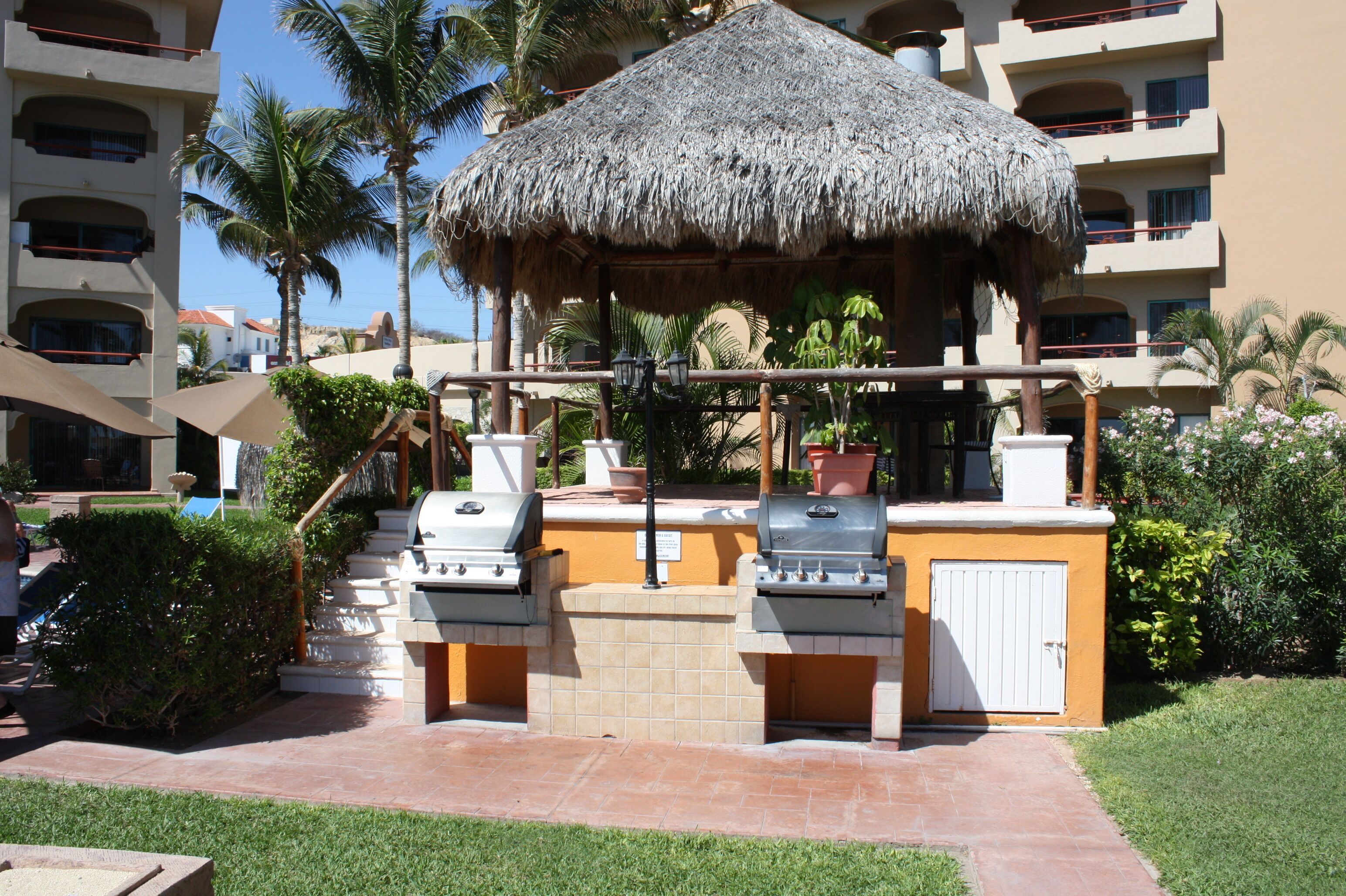 An outdoor barbecue area with two grills, a thatched-roof structure, and a poolside setting with palm trees and a building in the background.