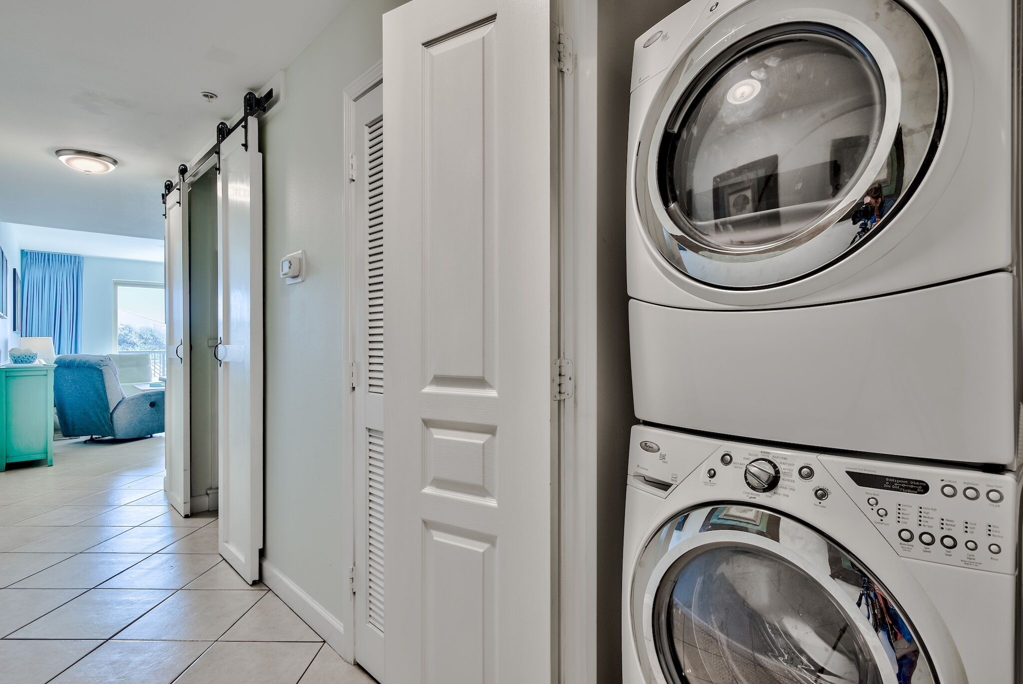 A laundry room with stacked washers and dryers, a teal side table, and a view of a living area with a blue chair and curtains.