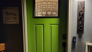 Une porte verte avec un paillasson, une pancarte indiquant « Bienvenue, amis », et une plaque de numéro au-dessus de la porte.