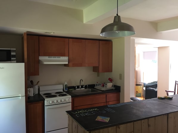 A kitchen with wooden cabinets, a white stove, a black countertop, and a chalkboard with 'Welcome First' written on it. - In town, river across the street, families welcome (Waitsfield)