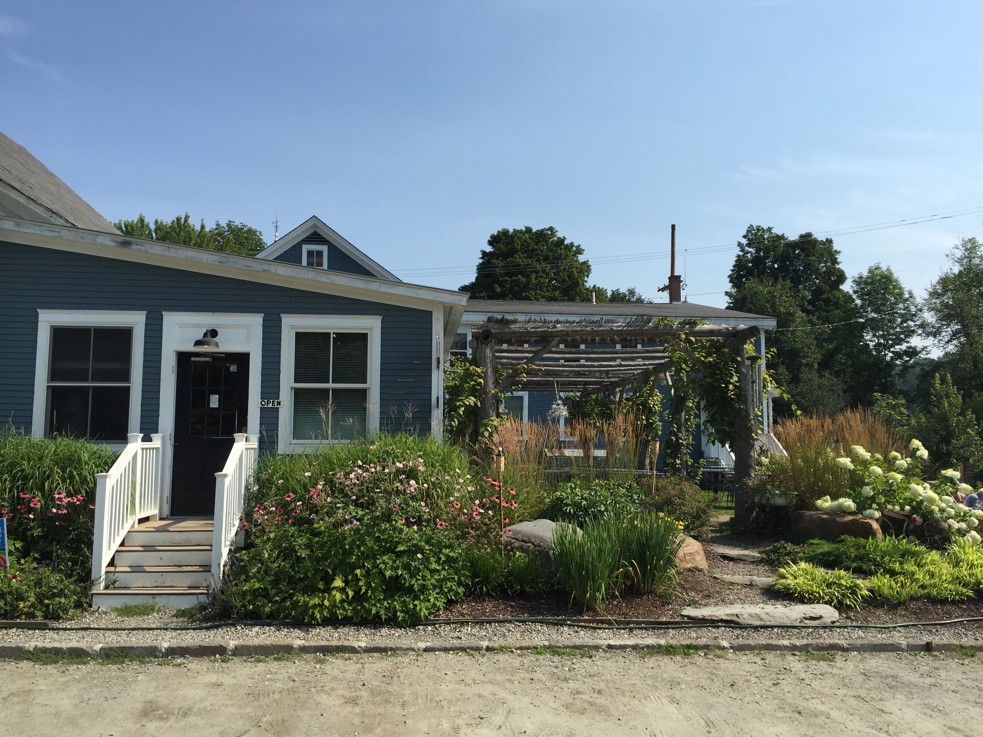 A two-story house with a front porch, a visible 'OPEN' sign, and a garden with various plants and flowers.