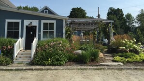 Et toetagers hus med en veranda, et synligt 'OPEN' skilt, samt en have med forskellige planter og blomster.