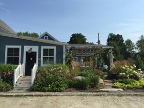 A two-story house with a front porch, a visible 'OPEN' sign, and a garden with various plants and flowers. - In town, river across the street, families welcome (Waitsfield)