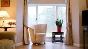Ein Wohnzimmer mit großem Fenster, einem beigen Sessel, einem kleinen Tisch mit Lampe und einer Topfpflanze.