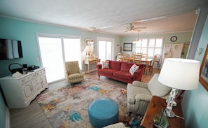 A living room with a sofa, ottoman, and chairs, a television, a dining table with chairs, and a ceiling fan. - Rideout's Hideout - Easy Beach Access - Sound Views - Family & Pet Friendly (Topsail Beach)
