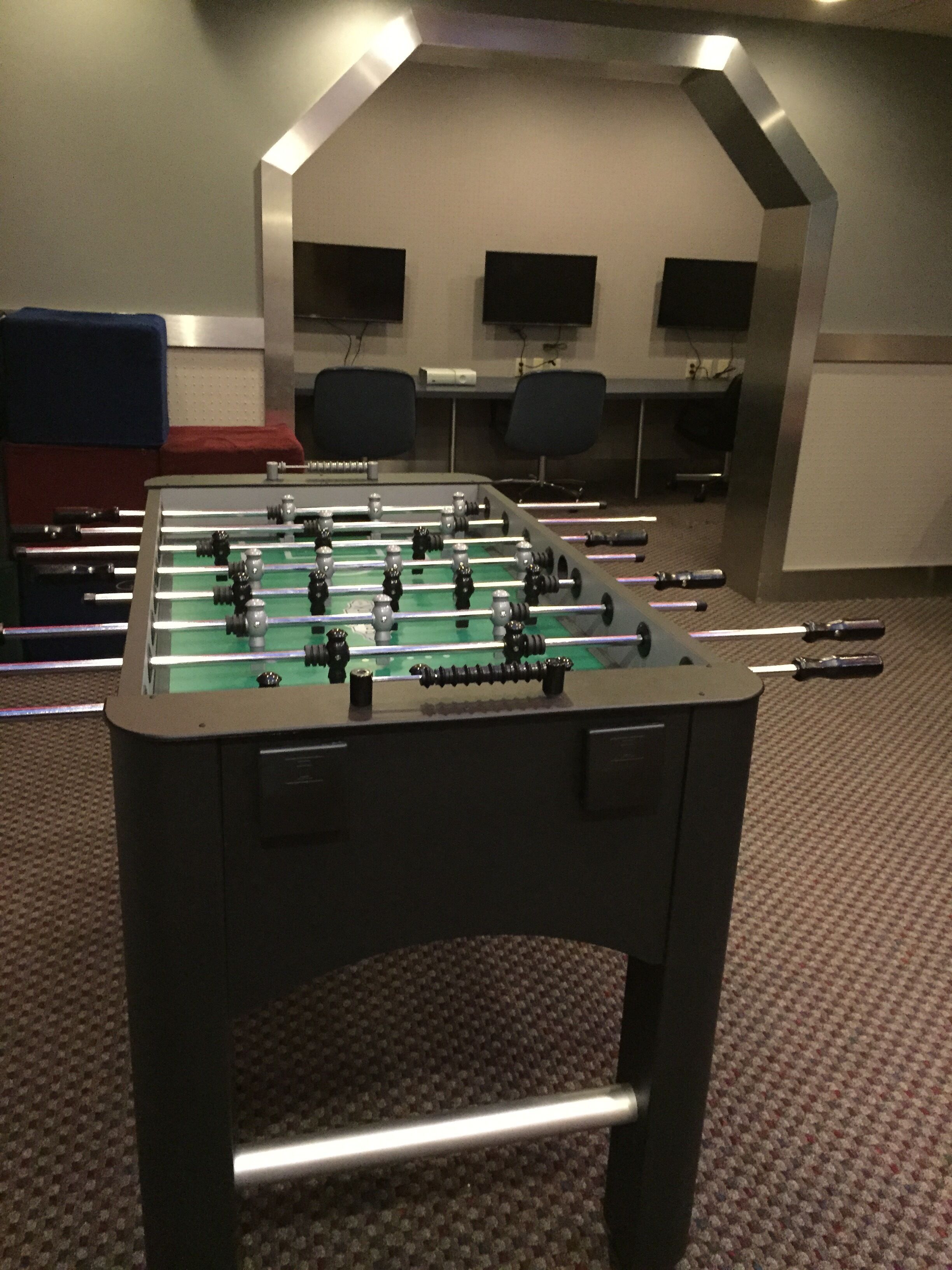 Una mesa de fútbol de mesa en una habitación con piso alfombrado, dos sillas y dos monitores en la pared.