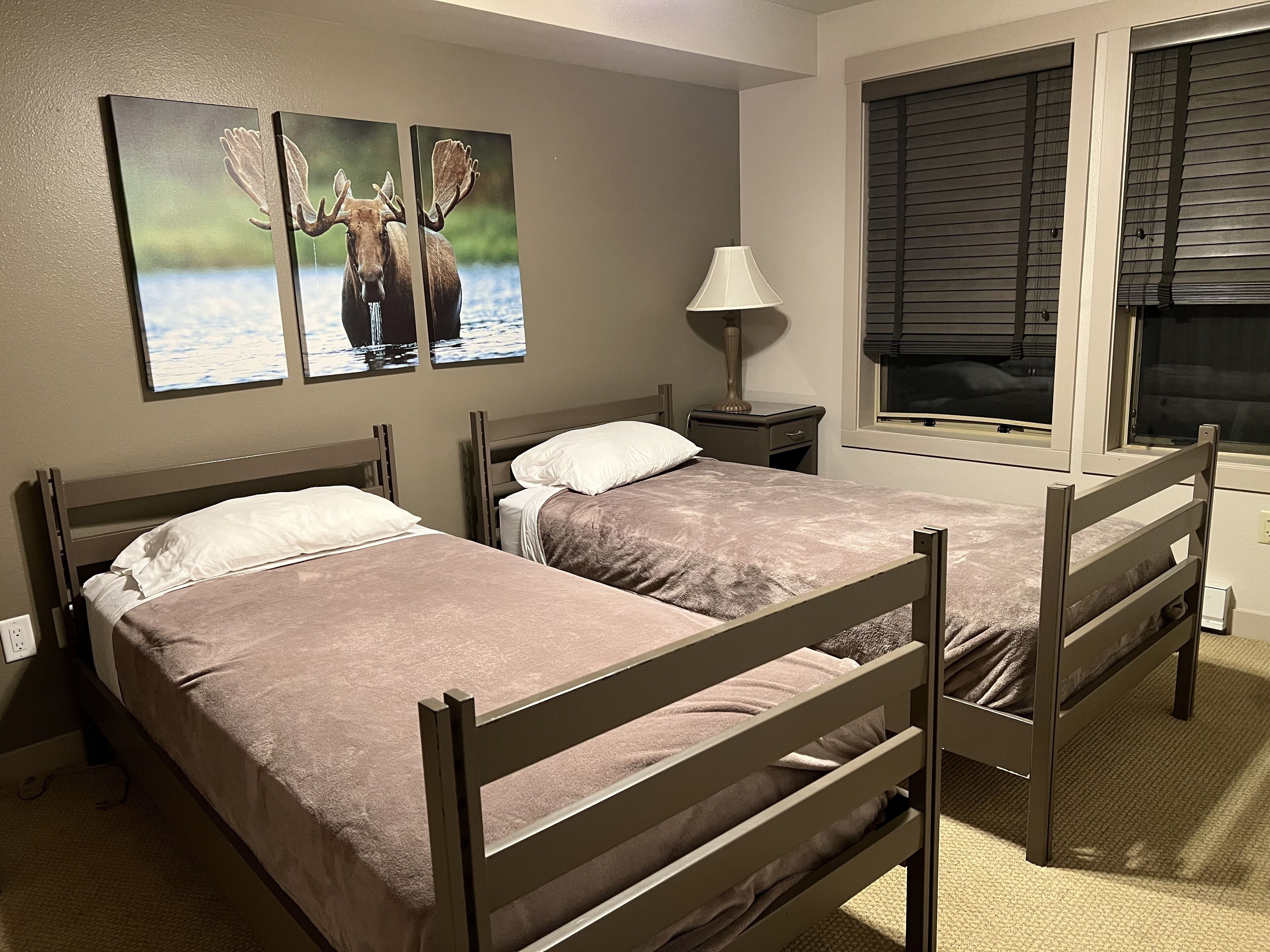 A room with two wooden beds, a nightstand with a lamp, a window with blinds, and two moose paintings on the wall.