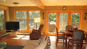 A living room with a sofa, a television, a dining table with chairs, and large windows with a view of trees. - Riverside 3 bedroom 2 bath near Baxter State Park (Millinocket)