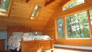 A wooden cabin bedroom with a bed, a nightstand, and large windows offering a view of the forest. - Riverside 3 bedroom 2 bath near Baxter State Park (Millinocket)