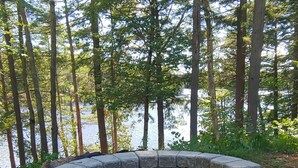 A circular stone fire pit surrounded by a forested area with tall trees and a body of water in the background. - Riverside 3 bedroom 2 bath near Baxter State Park (Millinocket)