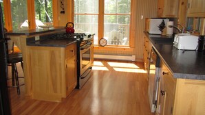 A kitchen with wooden cabinets, a stove, and a window overlooking a deck. - Riverside 3 bedroom 2 bath near Baxter State Park (Millinocket)