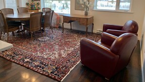 A dining area with a round table, wicker chairs, a red leather sofa, a patterned rug, and large windows with a snowy landscape view. - 2 Bedroom Apt RIVER & MOUNTAIN VIEWS , IN THE HEART OF WHITEHORSE - condo (Whitehorse)