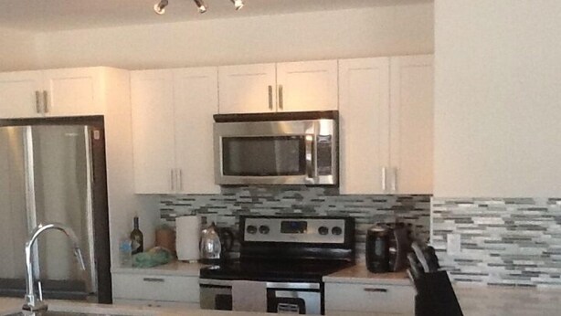 A modern kitchen with white cabinets, a stainless steel refrigerator, a microwave, a stove, and a bar area with two stools. - 2 Bedroom Apt RIVER & MOUNTAIN VIEWS , IN THE HEART OF WHITEHORSE - condo (Whitehorse)