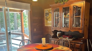 A dining area with a round table covered in a colorful tablecloth, surrounded by wooden chairs. A chandelier hangs above, and a wooden cabinet with glass doors is to the right.