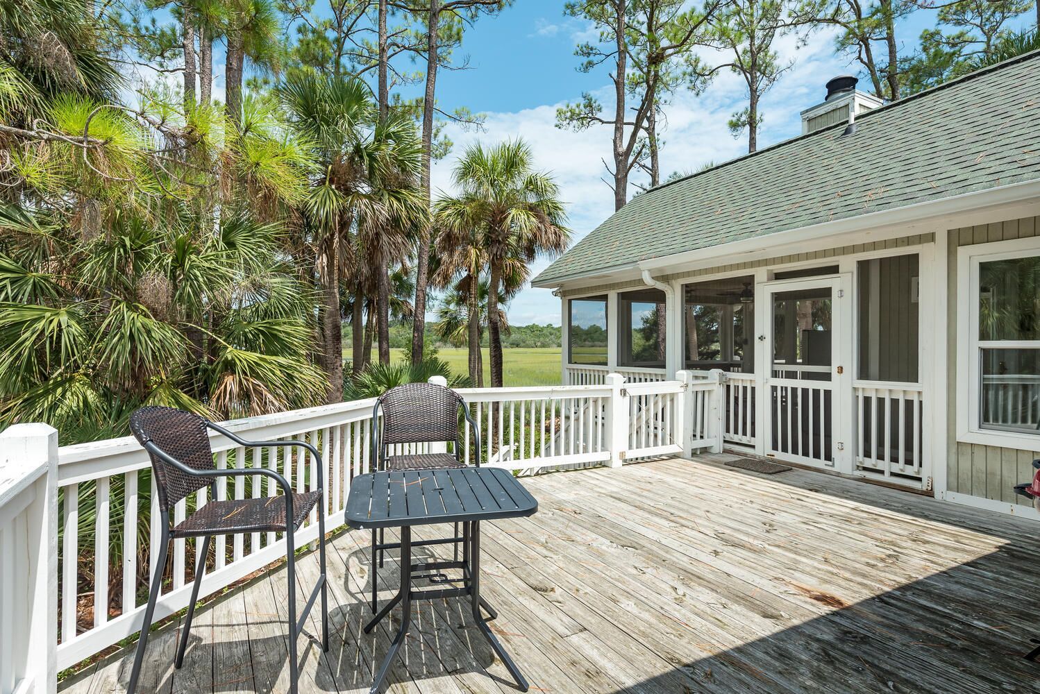 A wooden deck with a table and chairs, surrounded by white railings and palm trees.