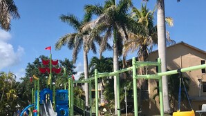 A playground with a slide and swings, surrounded by palm trees and a building in the background. - Florida Keys Townhouse For Rent 2/2 (Key Largo)