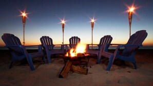 Quatro cadeiras Adirondack dispostas em volta de uma fogueira numa praia de areia ao pôr do sol.