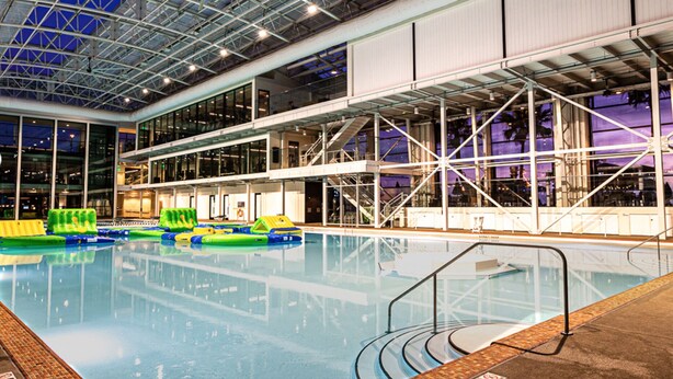 Piscina interior com água cristalina, rodeada por teto alto com vigas estruturais aparentes e painéis de vidro.