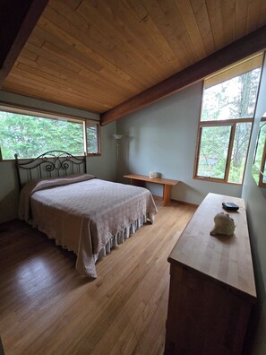 Une chambre avec un plafond en bois, un lit recouvert d’une couvre-lit à motifs, une table de chevet et une commode. Deux fenêtres laissent entrer la lumière naturelle et offrent une vue sur les arbres.