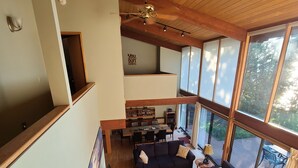 A spacious living area with a high ceiling, wooden beams, a dark sofa set, a patterned rug, and large windows offering a view of the outdoors.
