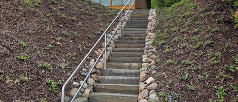 A set of stone steps leading up to a building with a metal railing, surrounded by trees and rocks.