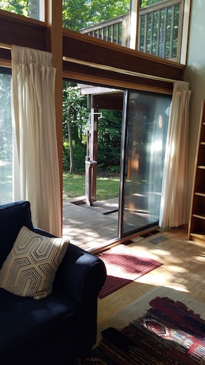 A room with a dark blue sofa, a patterned pillow, and a red and beige rug. There is a sliding glass door leading to an outdoor area with a wooden deck and greenery. - Private Lakefront Suttons Bay Home with Hot Tub, Newly Renovated Beach and A/C! (Suttons Bay)